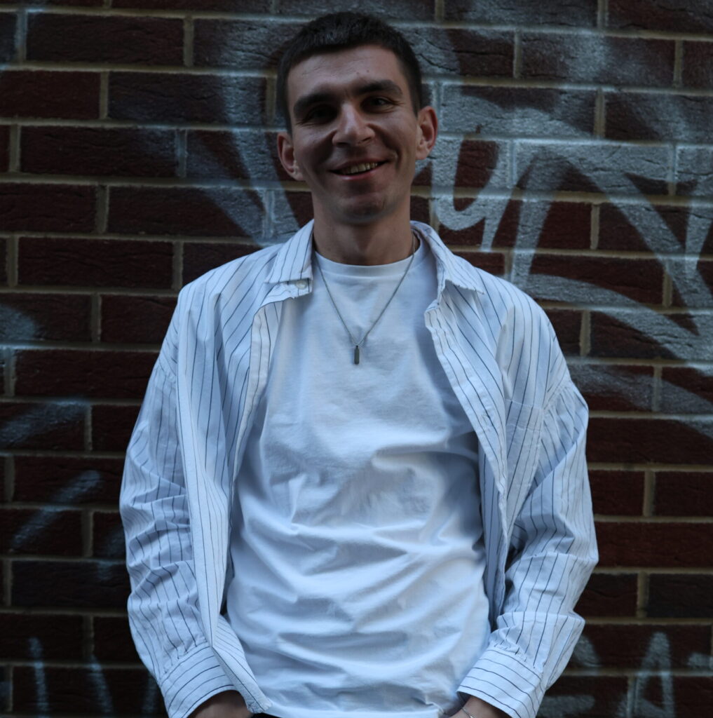 Portrait of Artem Nykyforov (ARNY), a Ukrainian music producer, vocal coach and vocalist, standing in front of a graffiti-covered brick wall in London, wearing a white t-shirt and striped shirt with a relaxed smile.
